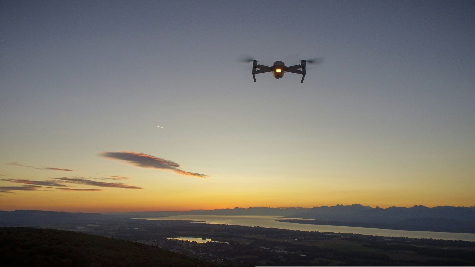 Drone with its lights on, flying at dusk above a valley and lake.